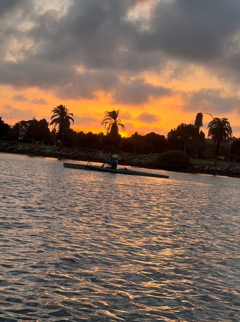 A paddler on an OC1, and in the background a cloudy sunset behind a dark shore lined with palm trees.